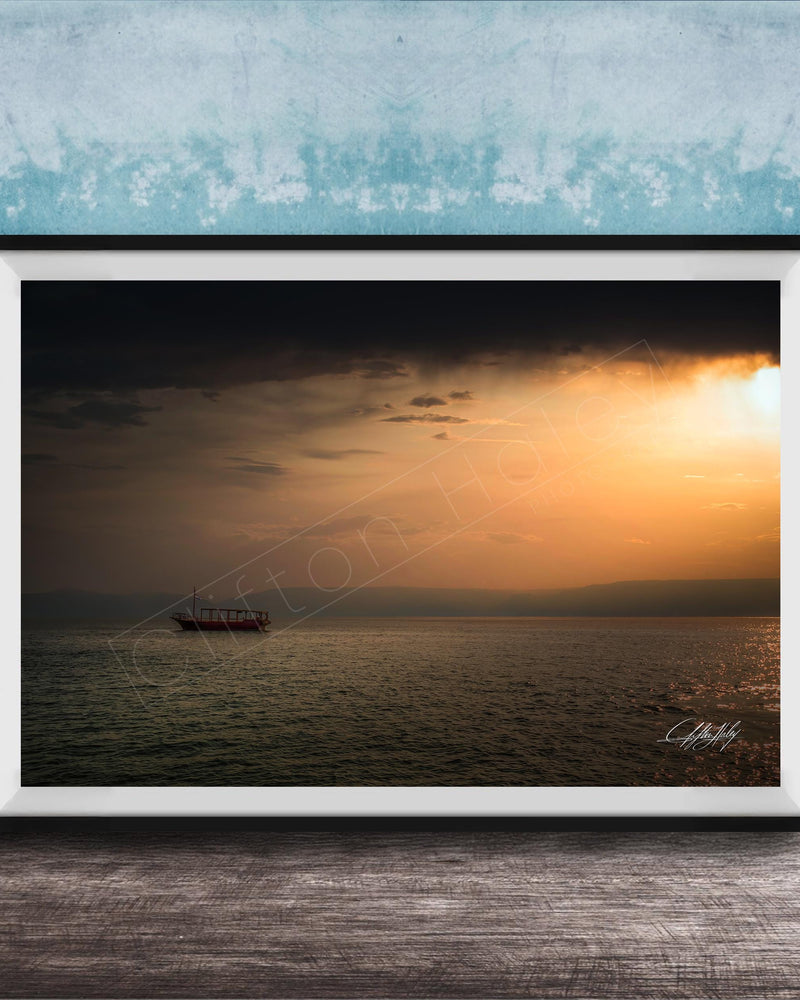 A framed photograph of a boat sailing on a calm body of water at sunset, with a blue sky and clouds in the background.