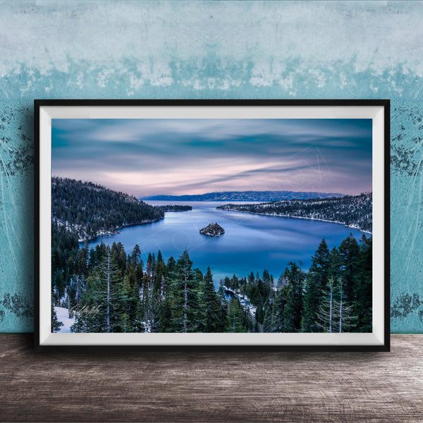 A framed photograph depicting a serene landscape with a lake surrounded by snow-covered trees and mountains in the distance, set against a blue sky with wispy clouds.