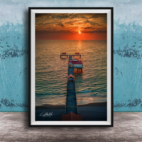A framed photograph depicting a serene sunset over a body of water, with a pier extending into the water and a small building on the pier.