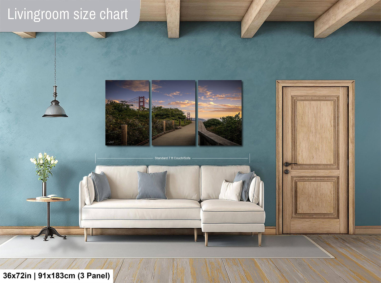 The image is a living room with a large wall art depicting a scenic view of the Golden Gate Bridge at sunset. The living room has a white sofa, a wooden door, and a wooden table with a vase of flowers. The wall art is approximately 36 inches wide and 15 inches tall, and it is a three-panel collage.