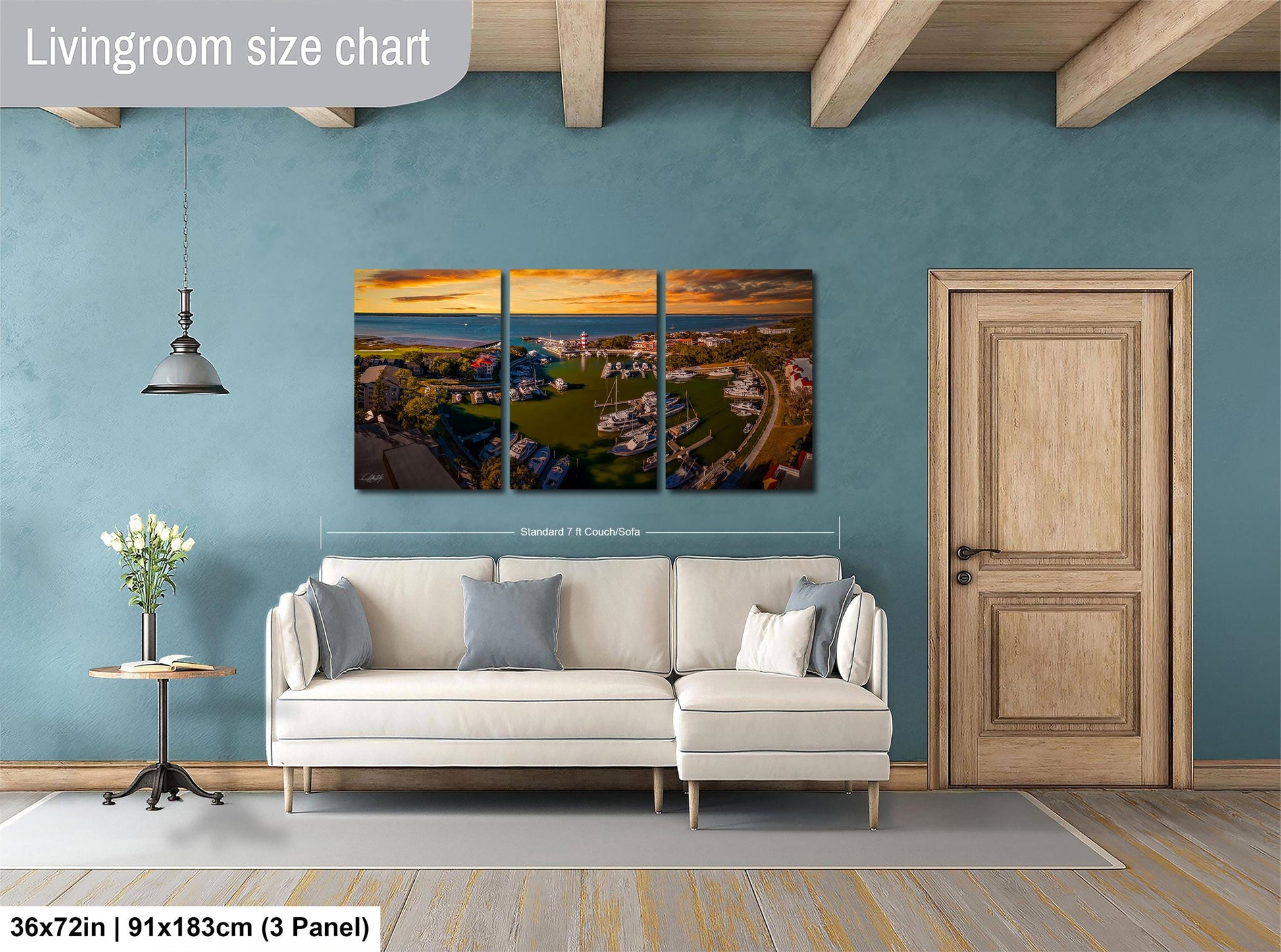 The image is a living room with a large, three-panel landscape painting hanging on the wall. The painting depicts a cityscape with buildings, roads, and a body of water. The room has a white sofa and a wooden door, and there is a vase of flowers on a table. The painting is approximately 36 inches wide and 91 inches tall, and it is a collage of three smaller images.