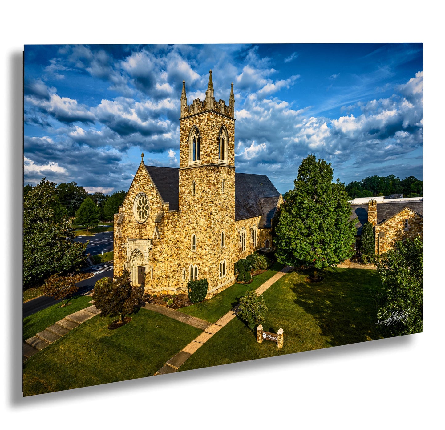 A stone church with a tall tower, surrounded by trees and a grassy lawn, set against a blue sky with clouds.