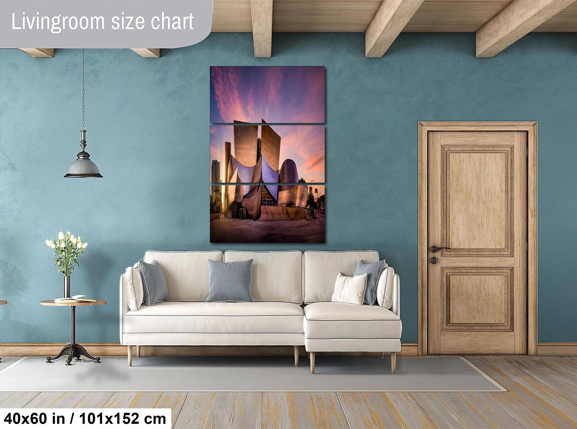 The image is a living room with a large, colorful painting of a building on the wall. The painting is divided into three sections, with the top section showing a sunset, the middle section depicting a city skyline, and the bottom section showing a building. The room has a white sofa, a wooden door, and a small table with a vase of flowers. The painting is approximately 40 inches wide and 101.5 inches tall, and the room has a wooden floor.