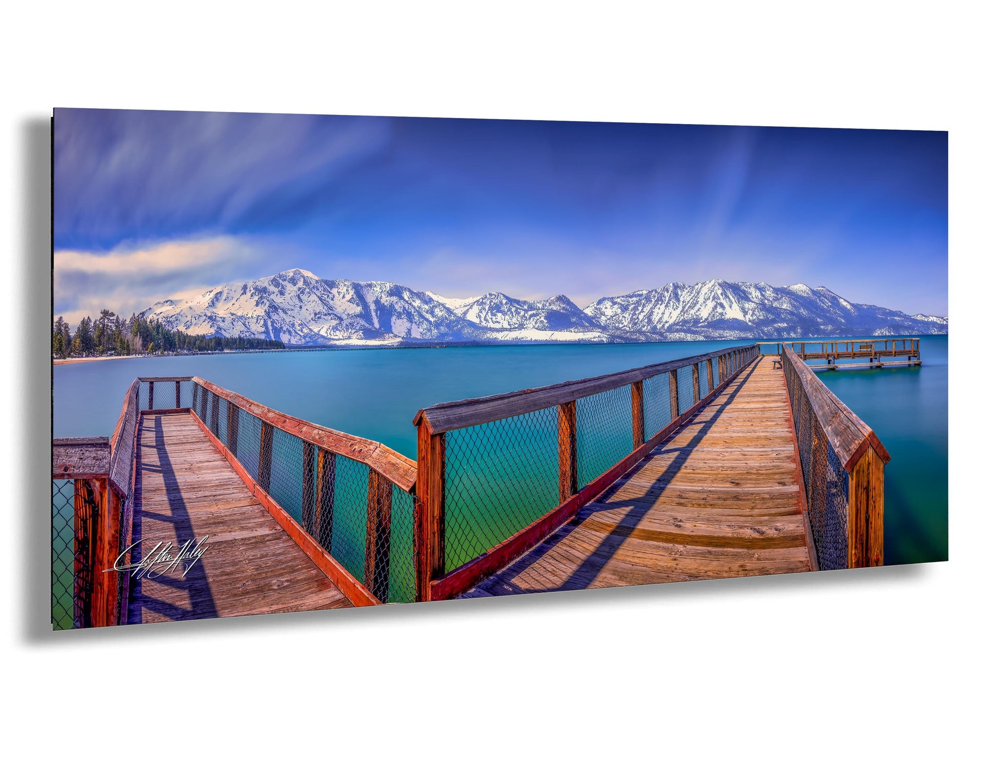 A wooden pier extends into a serene lake, with snow-capped mountains rising in the distance against a clear blue sky.
