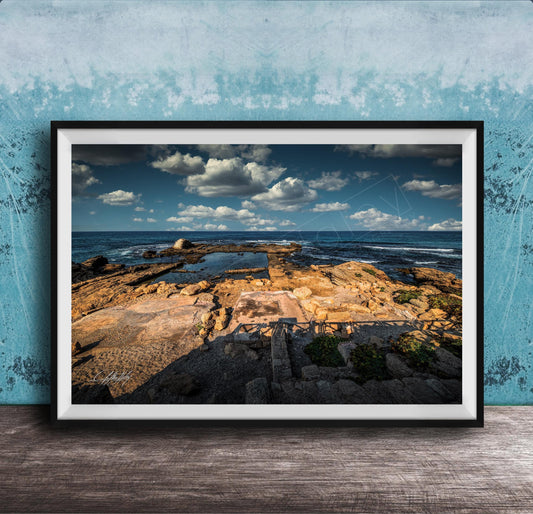 The image is a framed photograph of a rocky beach with a body of water in the background, set against a blue sky with clouds.