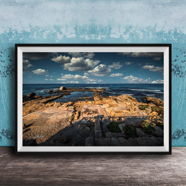 The image is a framed photograph of a rocky beach with a body of water in the background, set against a blue sky with clouds.