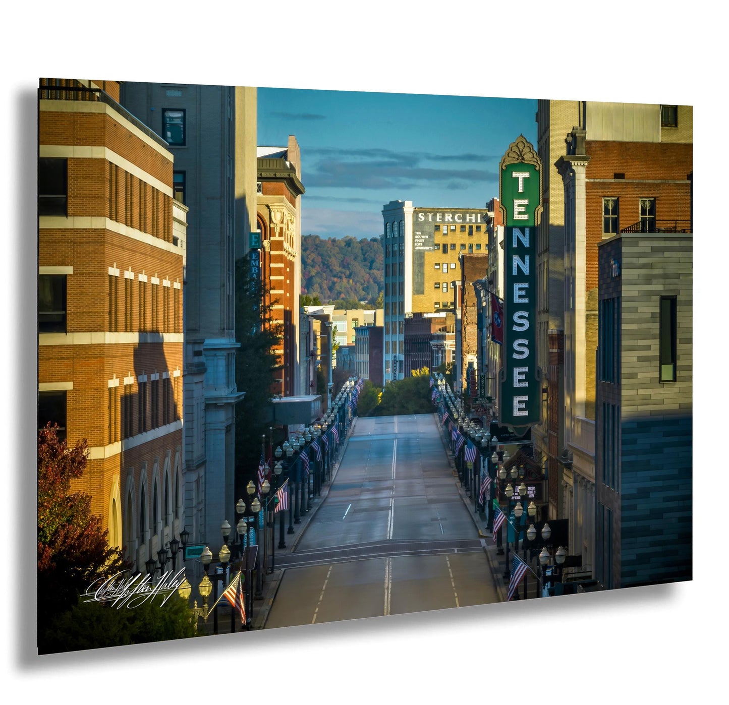 a city street lined with tall buildings, including a prominent sign for "Sterchi" and "TENNESSEE". The street is lined with flags and banners, creating a festive atmosphere. The sky is clear blue, and the sun is shining brightly, casting shadows and highlighting the architectural details of the buildings.