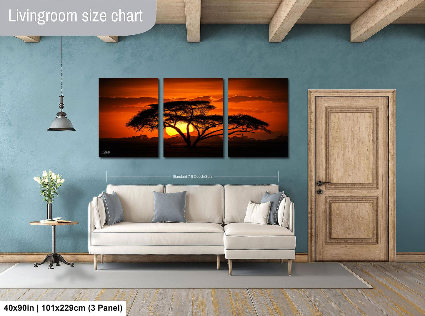 The image is a living room with a large wall art depicting a sunset over a tree. The wall art is divided into three panels, with the largest panel spanning approximately 40x50 inches (101x129 cm) and the smallest panel being smaller. The room also includes a white sofa, a wooden door, and a small table with a vase of flowers.