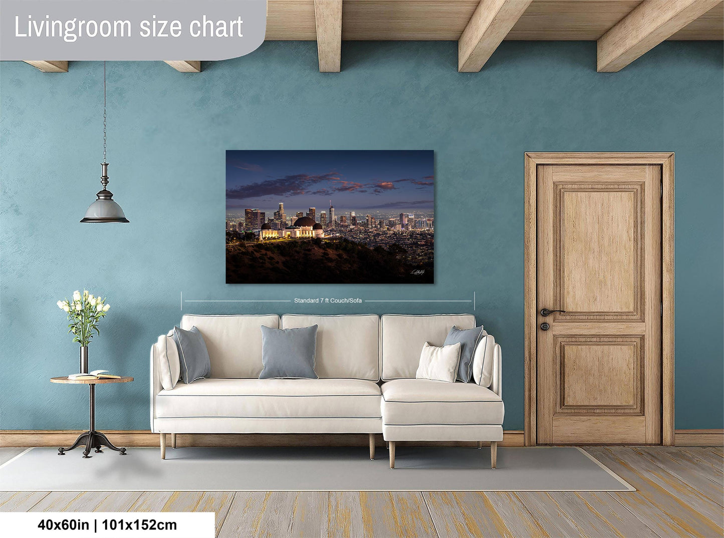 a living room with a large painting of a city skyline hanging on the wall. The painting is framed and measures 40 inches by 101.5 centimeters. The room has a white sofa, a wooden door, and a small table with a vase of flowers.