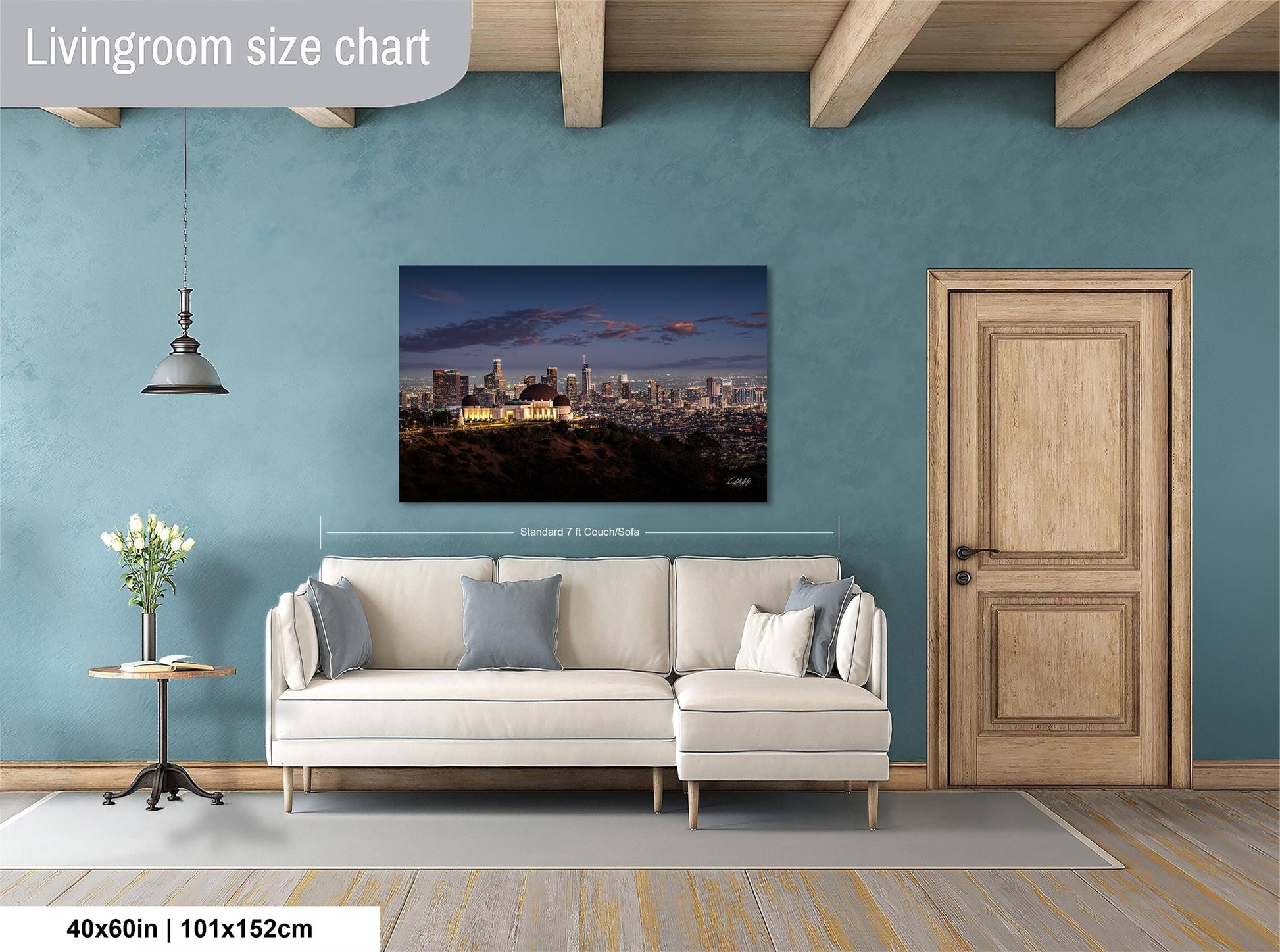 a living room with a large painting of a city skyline hanging on the wall. The painting is framed and measures 40 inches by 101.5 centimeters. The room has a white sofa, a wooden door, and a small table with a vase of flowers.