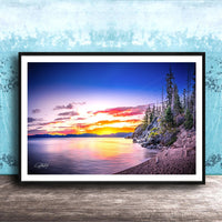 A framed photograph depicting a serene landscape with a calm lake reflecting the vibrant colors of a sunset sky, surrounded by trees and mountains.