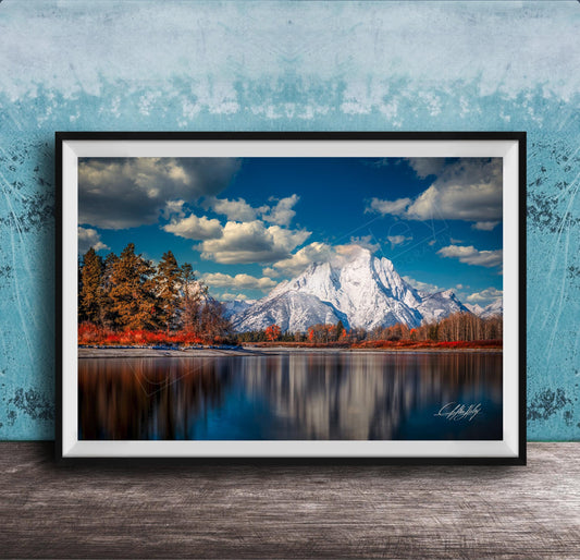 A framed photograph depicting a serene mountain landscape with a calm lake reflecting the surrounding scenery, including trees and mountains in the background.