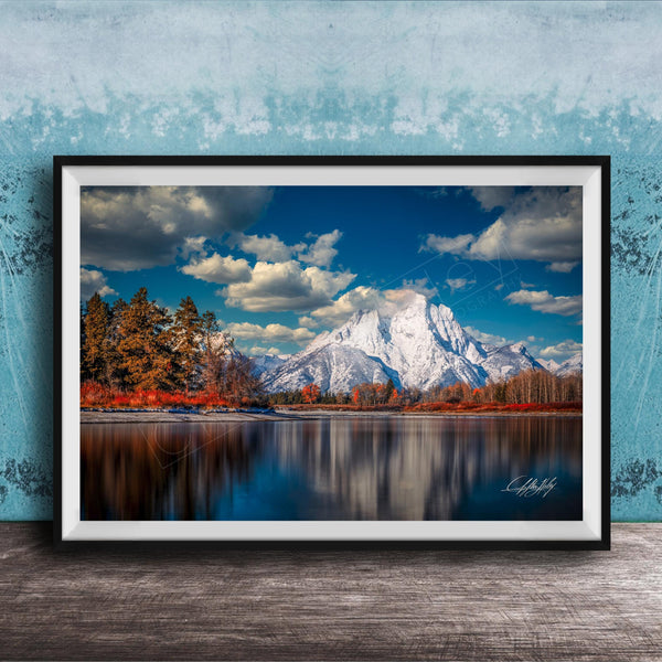 A framed photograph depicting a serene mountain landscape with a calm lake reflecting the surrounding scenery, including trees and mountains in the background.