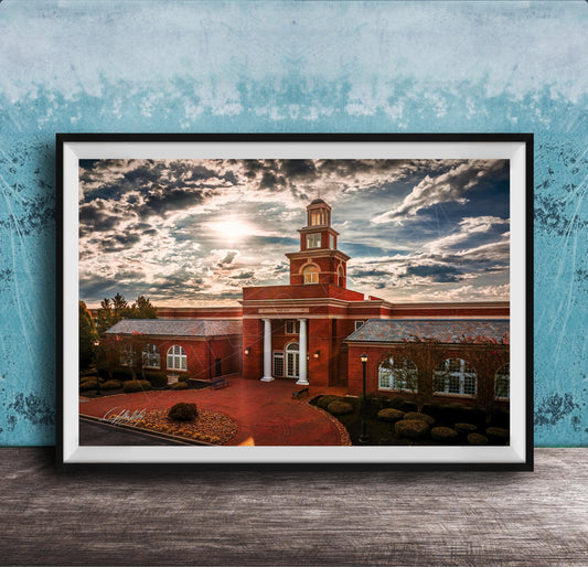 A framed photograph of a red brick building with a tower, set against a cloudy sky at sunset.