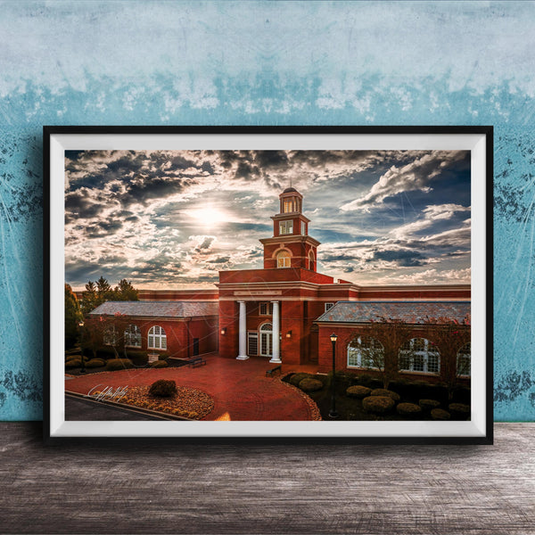 A framed photograph of a red brick building with a tower, set against a cloudy sky at sunset.
