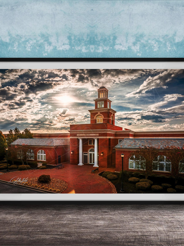 A framed photograph of a red brick building with a tower, set against a cloudy sky at sunset.