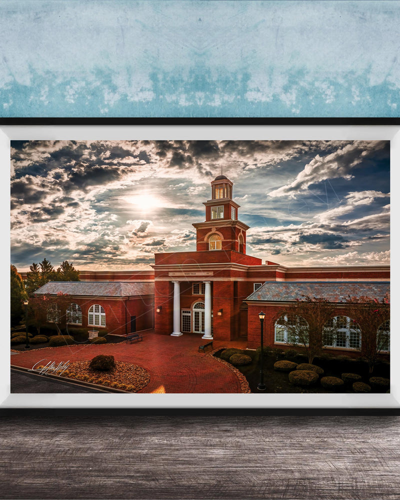 A framed photograph of a red brick building with a tower, set against a cloudy sky at sunset.