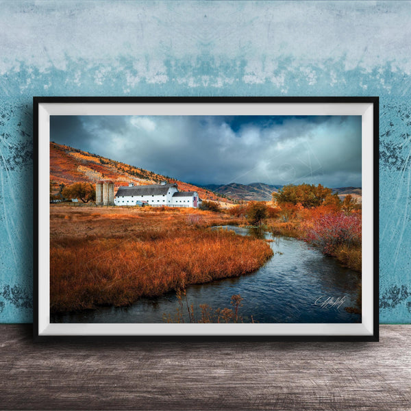 a picturesque rural landscape with a small white building nestled among the rolling hills and lush greenery. The scene is framed and displayed on a wooden surface, with a blue wall in the background.