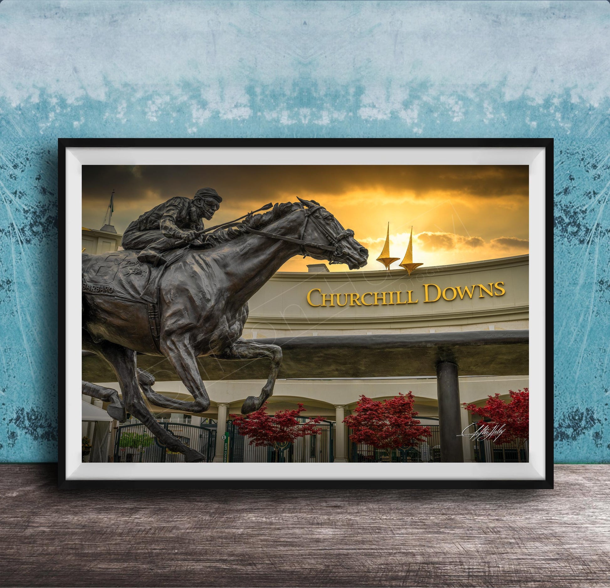 A bronze statue of a jockey riding a horse, with the words "Churchill Downs" displayed above it, against a backdrop of a sunset sky and sailboats.