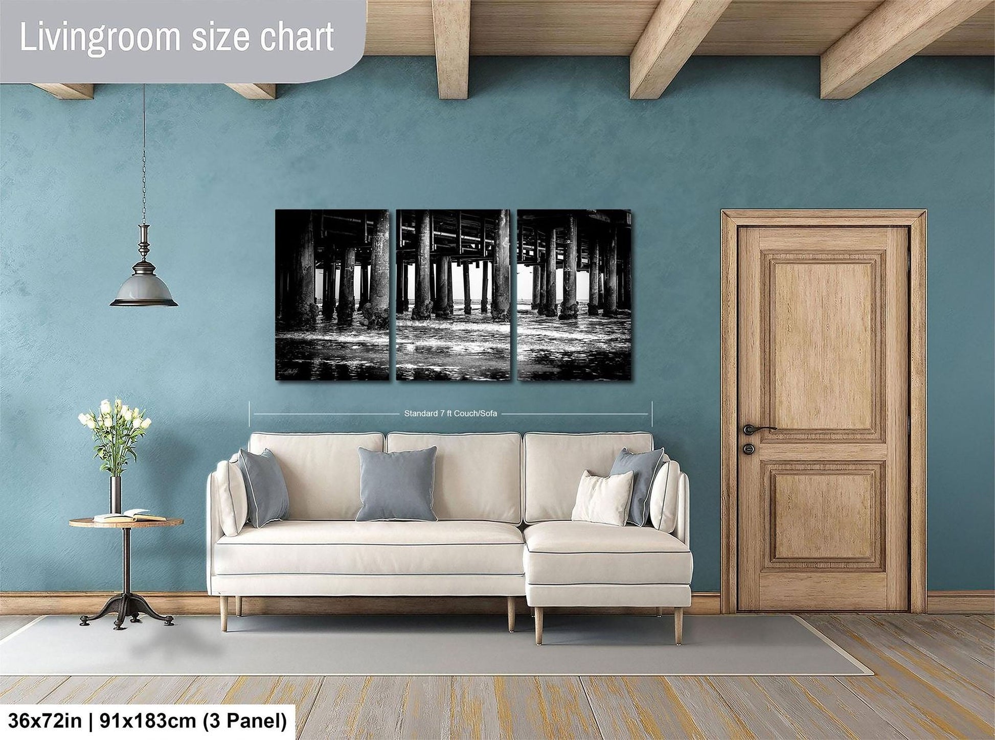 The image is a living room with a large white sofa, a wooden door, and a wall with a black and white photograph of a bridge. The room has a blue wall and a wooden floor.
