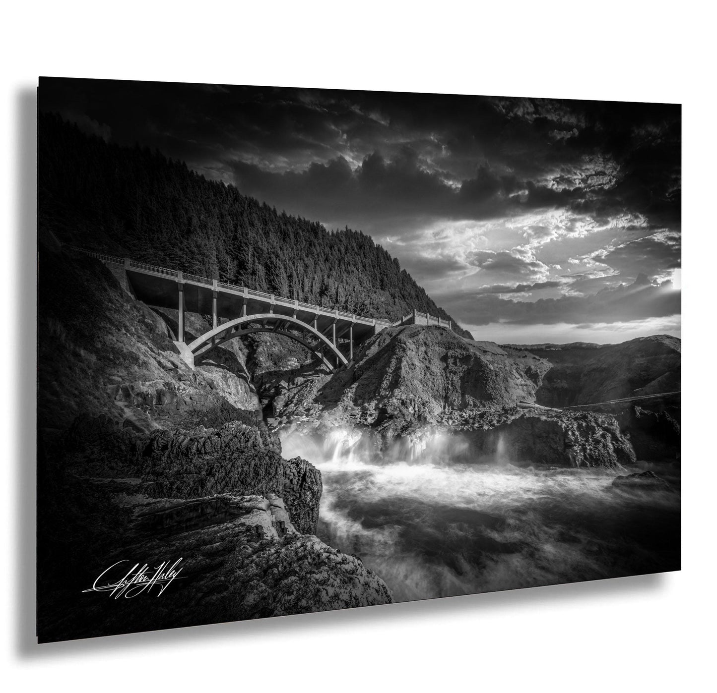 A dramatic black and white landscape photograph depicting a bridge spanning over a rushing river, with a dramatic sky and clouds in the background.