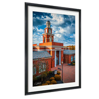 A framed photograph of a red brick building with a prominent clock tower, set against a blue sky with clouds.