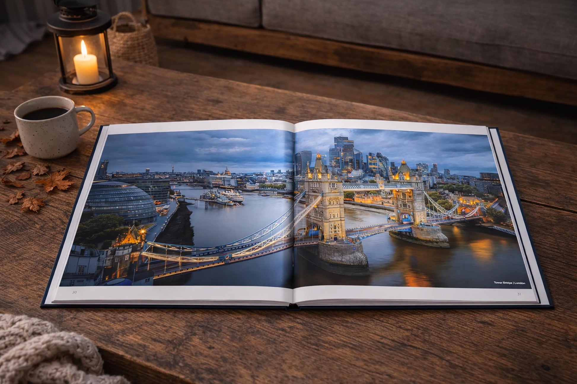An open book displaying a scenic view of London, with the iconic Tower Bridge and other cityscape visible. The book is placed on a wooden surface, with a lit candle in the background and some fallen leaves nearby.