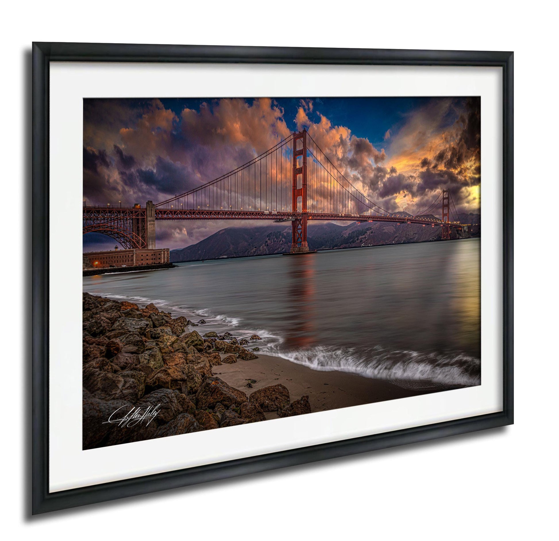 a picturesque scene of the Golden Gate Bridge in San Francisco, California, with its iconic red towers and suspension cables spanning the bay. The bridge is set against a dramatic sky filled with clouds and vibrant colors, creating a stunning backdrop for the serene waters of the bay. The photograph is framed and displayed on a wall, showcasing the beauty of this iconic landmark.