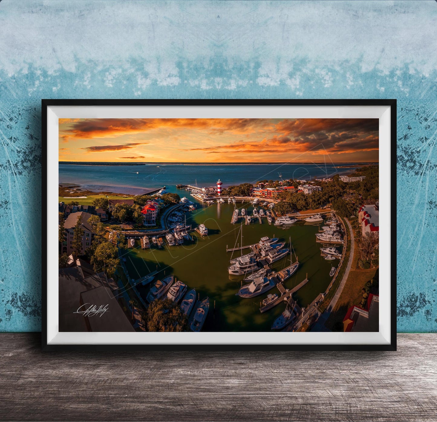 The image is a framed aerial view of a harbor with boats docked, set against a backdrop of a colorful sunset sky.