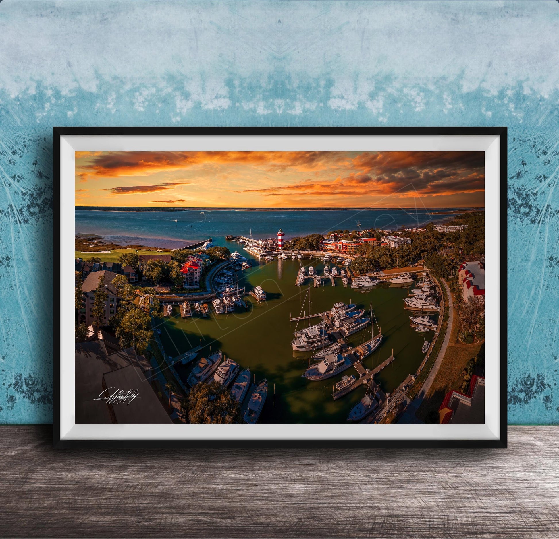 The image is a framed aerial view of a harbor with boats docked, set against a backdrop of a colorful sunset sky.