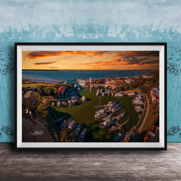 The image is a framed aerial view of a harbor with boats docked, set against a backdrop of a colorful sunset sky.