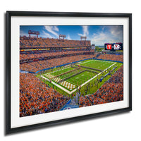 A framed image depicting a crowded football stadium with a large field, a scoreboard, and a large crowd of spectators.