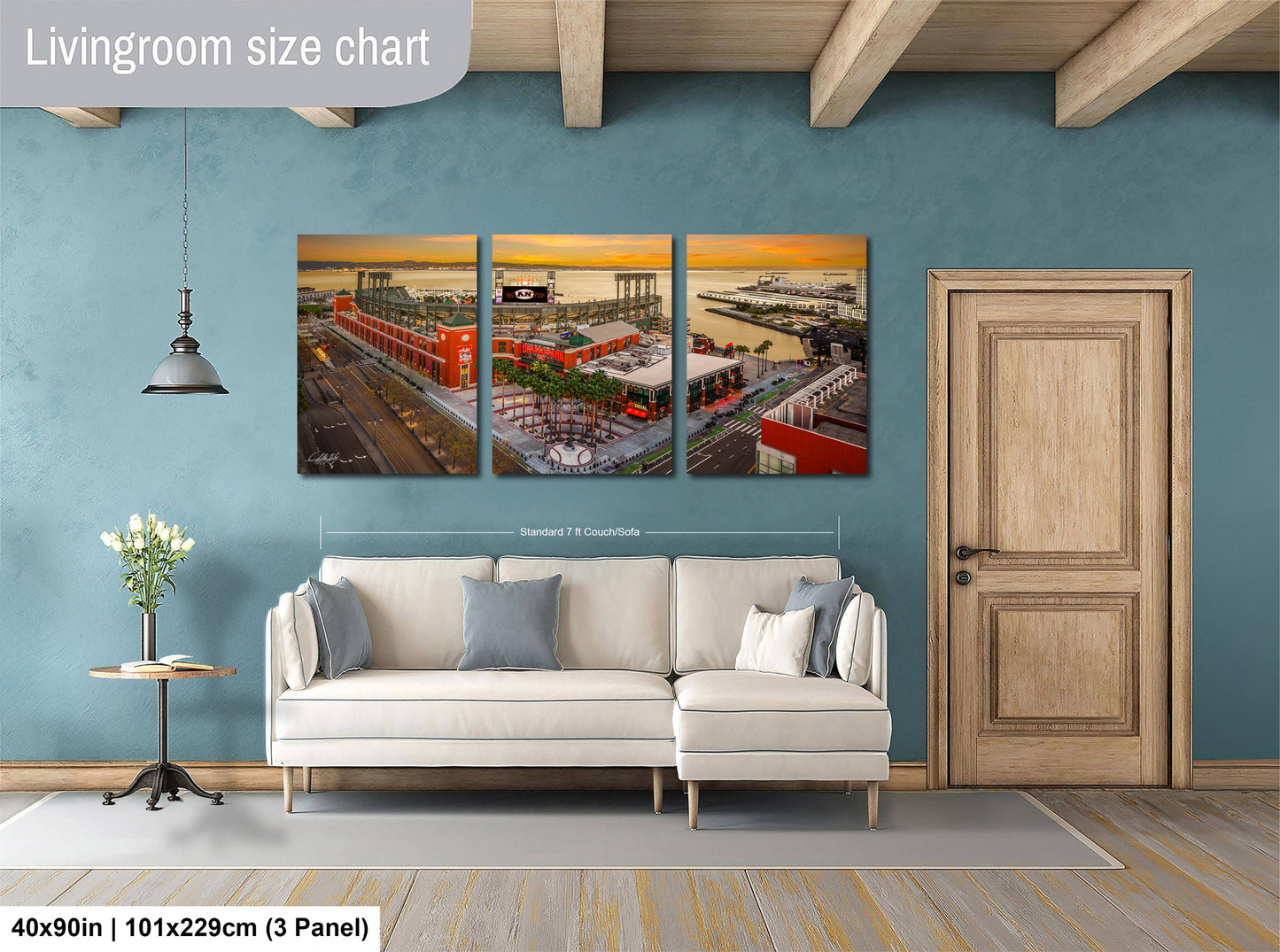 The image is a living room with a large white sofa and a wooden door. Above the sofa, there is a large wall art collage depicting a cityscape with buildings, roads, and a sunset. The collage is divided into three panels, each showing different aspects of the cityscape. The wall art is approximately 40 inches wide and 101.29 cm tall, with a 3-panel arrangement.