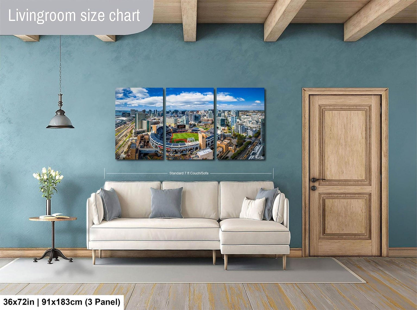The image is a living room with a large wall art depicting a cityscape. The wall art is a collage of three panels, each showing a different part of the city. The living room has a white sofa and a wooden door, and there is a potted plant and a vase on the floor. The wall art is approximately 36 inches wide and 9 inches tall.