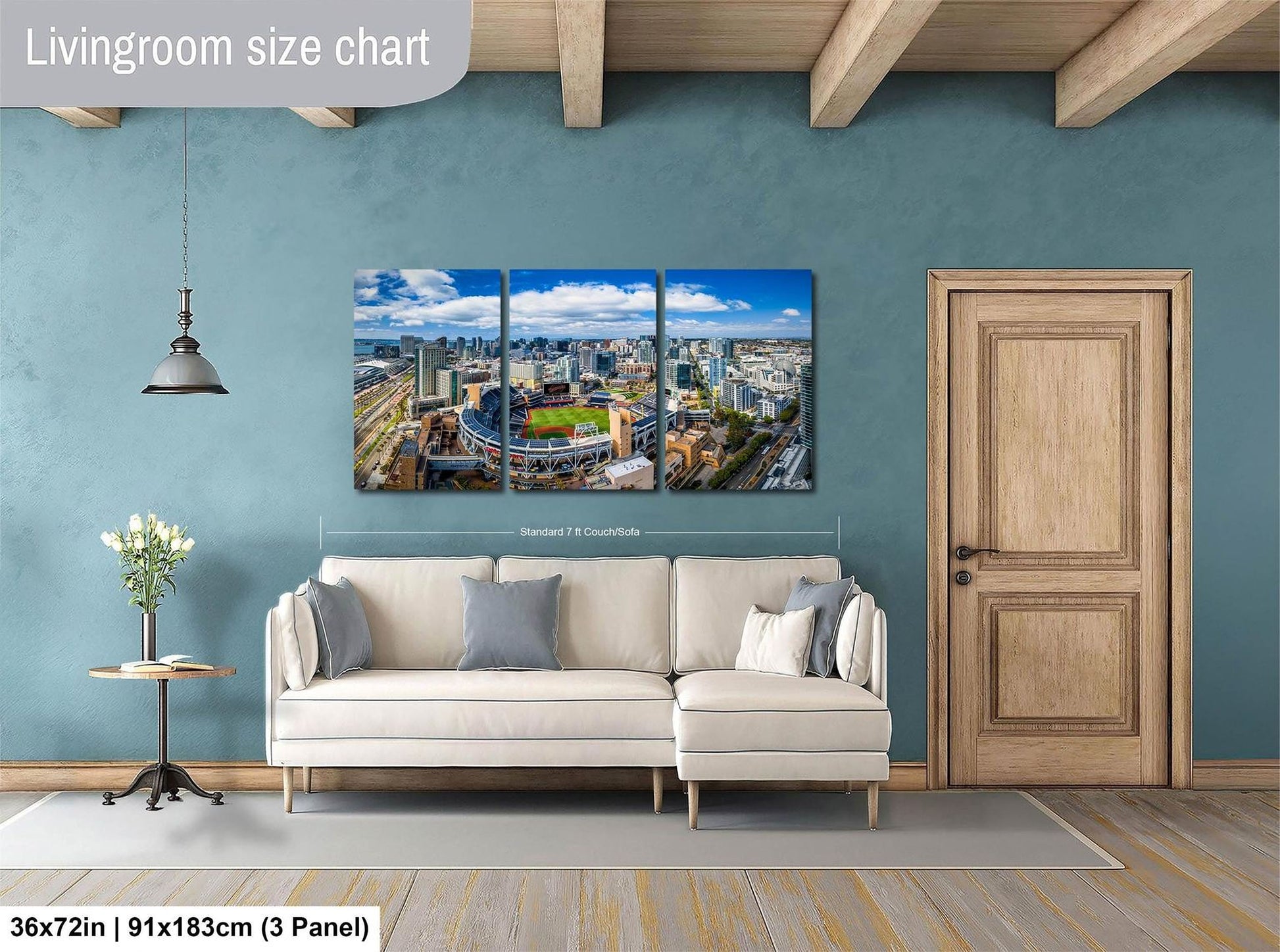 The image is a living room with a large wall art depicting a cityscape. The wall art is a collage of three panels, each showing a different part of the city. The living room has a white sofa and a wooden door, and there is a potted plant and a vase on the floor. The wall art is approximately 36 inches wide and 9 inches tall.