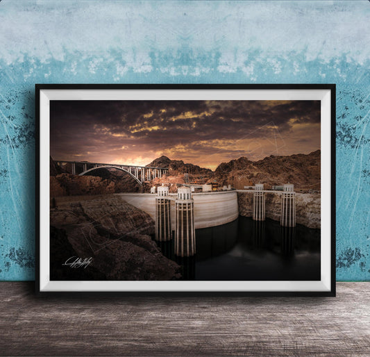 A framed photograph depicting a large dam with a bridge spanning across it, set against a dramatic sky with clouds and a sunset.