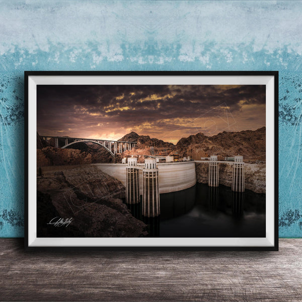 A framed photograph depicting a large dam with a bridge spanning across it, set against a dramatic sky with clouds and a sunset.