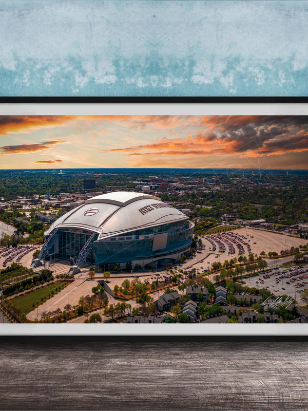 The image is a framed photograph of a large stadium with a curved roof, set against a backdrop of a city skyline and a colorful sunset sky.