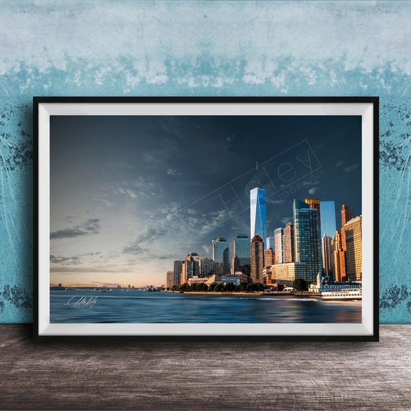 A framed photograph of the New York City skyline, with the One World Trade Center prominently visible, set against a blue sky with clouds.