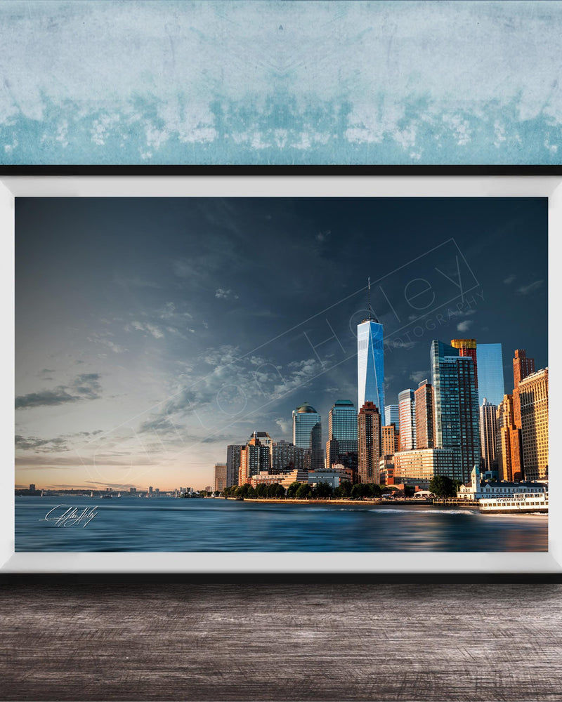A framed photograph of the New York City skyline, with the One World Trade Center prominently visible, set against a blue sky with clouds.