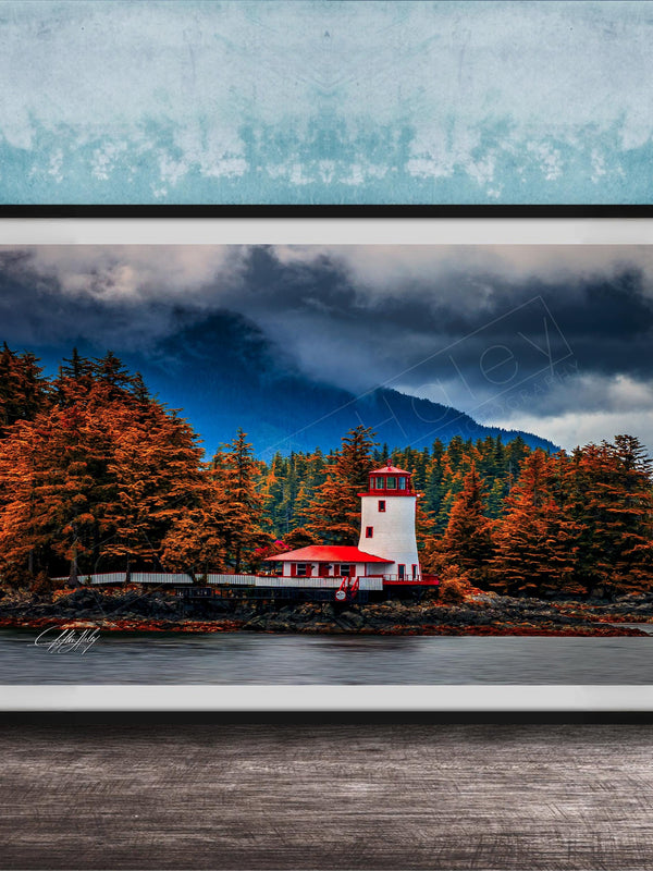 A framed photograph depicting a lighthouse surrounded by a forest with autumn-colored trees, set against a cloudy sky.