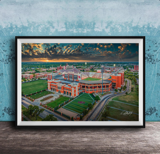 The image is a framed aerial view of a large sports stadium with a large field, surrounded by surrounding buildings and a cloudy sky.