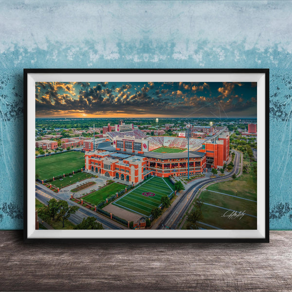 The image is a framed aerial view of a large sports stadium with a large field, surrounded by surrounding buildings and a cloudy sky.