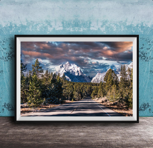 A framed photograph depicting a scenic mountain landscape with a winding road surrounded by pine trees and snow-capped peaks under a dramatic cloudy sky.