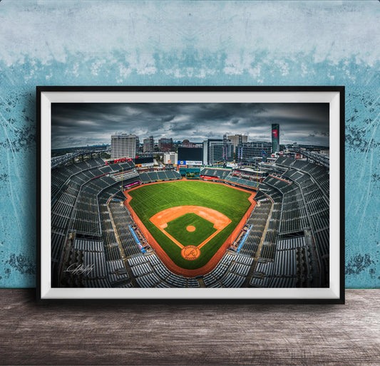 A framed photograph of a baseball stadium with a large field, surrounded by buildings and a cloudy sky.