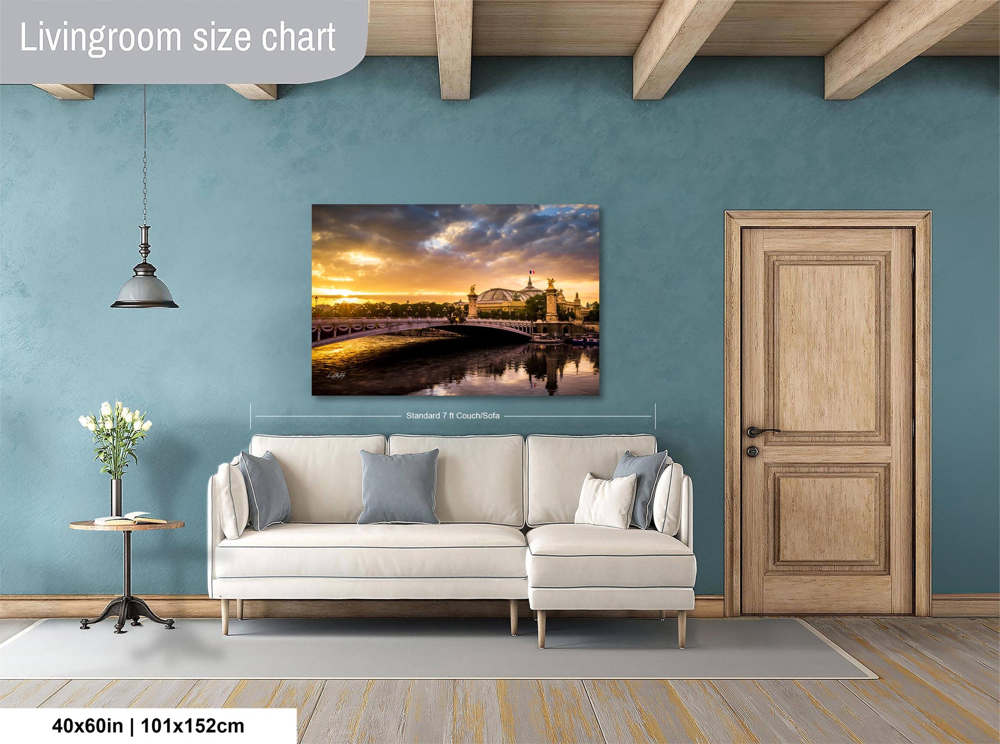 a living room with a large wall painting depicting a scenic view of a bridge and a sunset over a body of water. The painting is framed and hangs above a white sofa, which is positioned in front of a wooden door. The room has a wooden floor and a small table with a vase of flowers. The wall is painted in a light blue color, and the overall layout and design of the room create a cozy and inviting atmosphere.