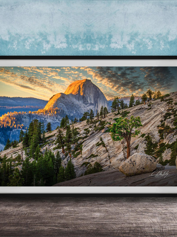 The image is a framed photograph depicting a mountainous landscape with a large mountain peak in the background, surrounded by trees and rocky terrain. The sky is filled with warm, golden light, creating a picturesque and serene atmosphere.