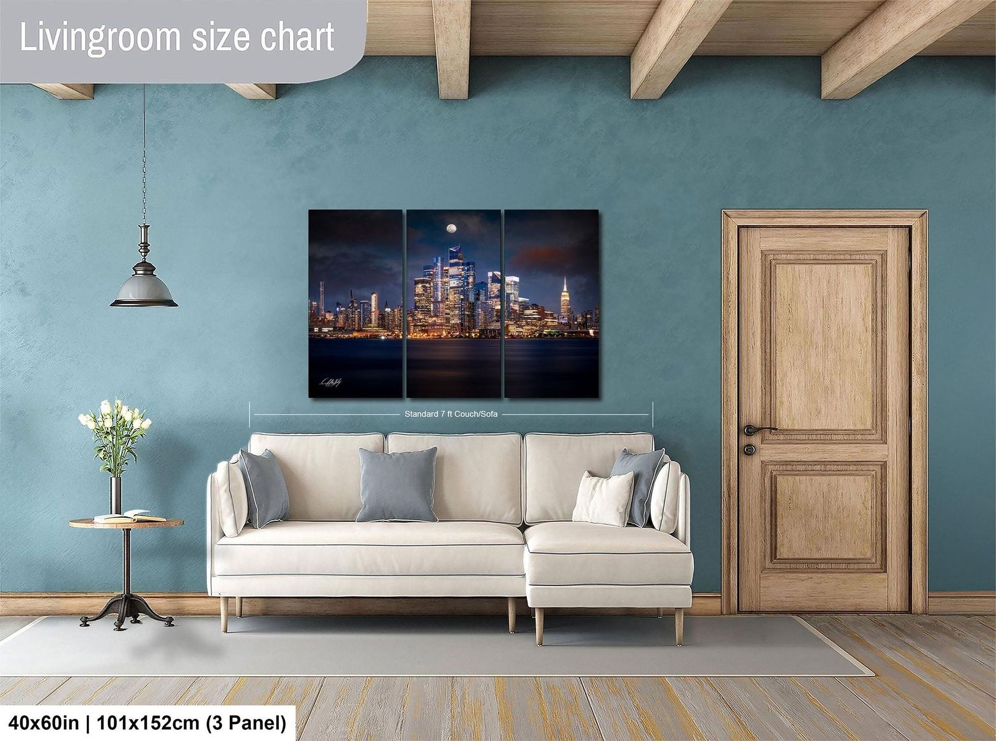 The image is a living room with a large, three-panel wall art depicting a city skyline at night. The wall art is mounted above a white sofa, and there is a wooden door and a small table with a vase of flowers nearby. The living room has a blue wall and wooden flooring.