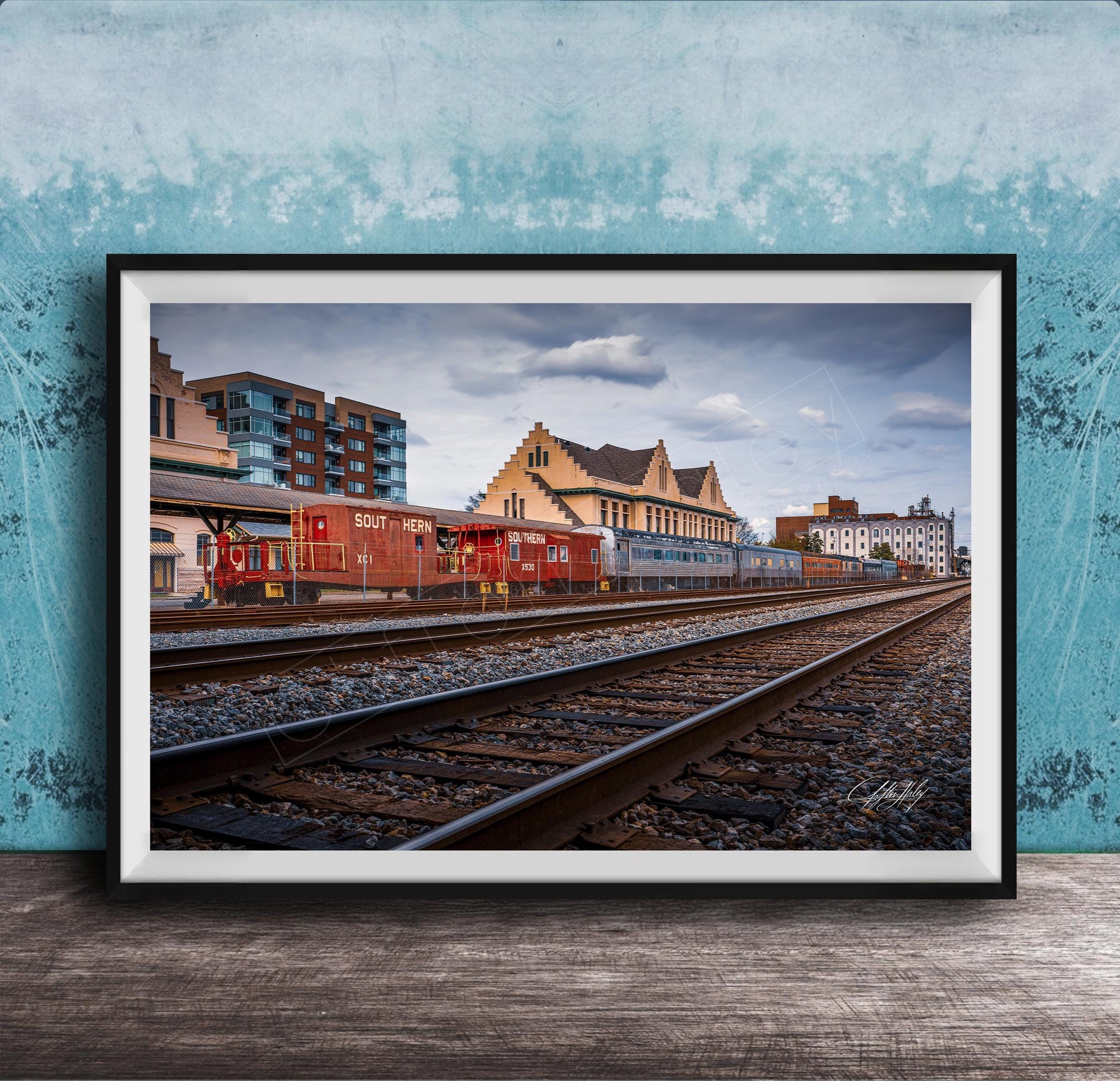 A framed photograph of a train on railroad tracks, with buildings in the background, set against a blue wall.