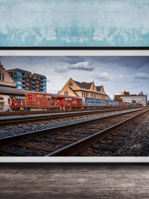 A framed photograph of a train on railroad tracks, with buildings in the background, set against a blue wall.
